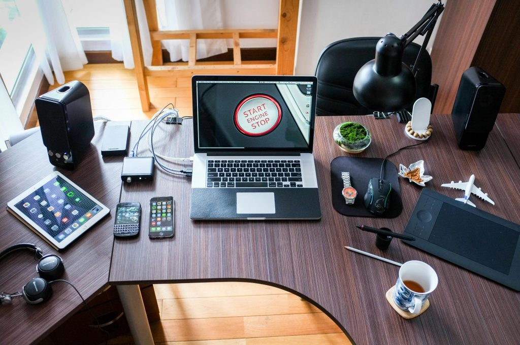 A modern home office setup featuring a MacBook with "Start Engine Stop" on the screen, surrounded by various gadgets including an iPad, smartphones, external hard drives, headphones, a digital drawing tablet, mouse, wristwatch, speakers, and decorative items on a U-shaped wooden desk.