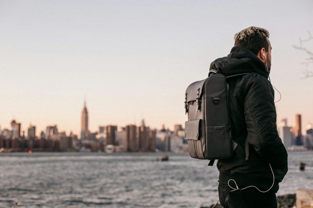 A man wearing a black jacket and backpack stands near the waterfront, facing the New York City skyline at sunset, with earphones connected to a device in his pocket.